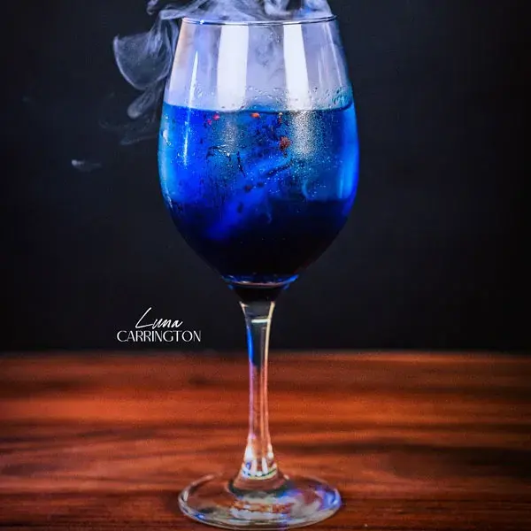Cóctel de mixología artística Luna Carrington en copa de vino, con un líquido azul vibrante y místico que desprende un efecto de humo denso, presentado sobre una mesa de madera oscura.