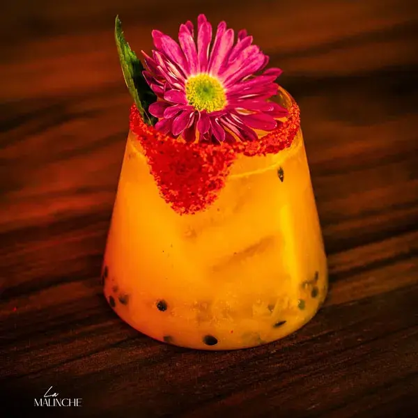 Bebida premium Malinche en vaso corto con rodaja de limón sobre fondo oscuro con flores, diseño exclusivo de coctelería de autor.