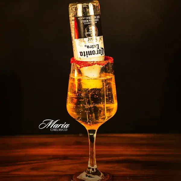 Cóctel de cerveza Maria Chelasco de mixología artística servido en copa de vino con borde escarchado de chile y una botella de Coronita Extra invertida adentro.