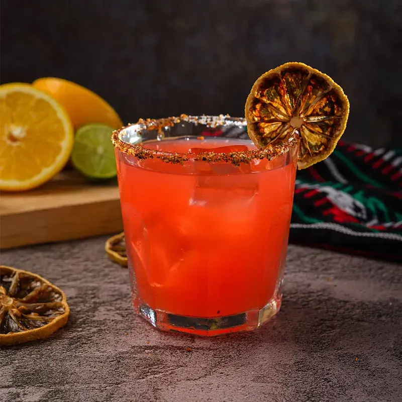 Cóctel de autor color rojo con hielo, escarchado artesanal de chile y rodaja de cítrico deshidratado para mixología artística.