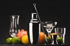 Coctelera de acero, jigger, cuchara de bar y frutas frescas sobre fondo oscuro, utensilios para mixología artística.