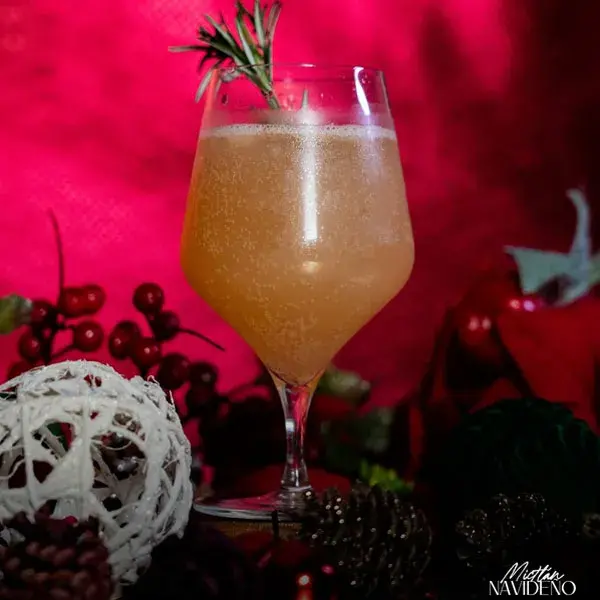 Cóctel festivo Mictlán Navideño de mixología artística, servido en copa con una ramita de romero sobre un fondo rojo con esferas y piñas navideñas.