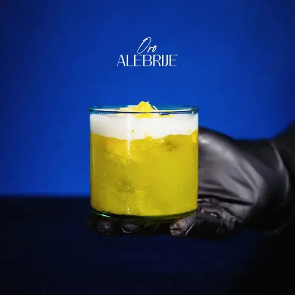 Cóctel de mixología artística Oro Alebrije en vaso corto con líquido amarillo brillante y espuma blanca, sostenido por una mano con guante negro sobre un fondo azul.